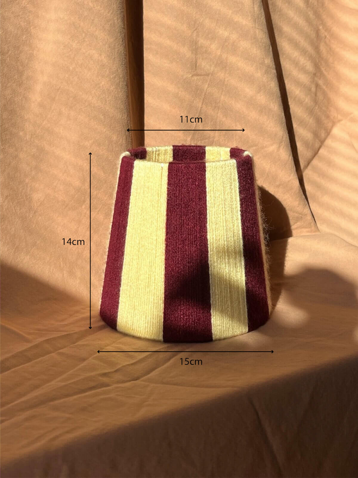 A small woven string lampshade with vertical stripes in deep burgundy and butter yellow, set against a draped beige fabric backdrop with strong sunlight and shadows with measurments.
