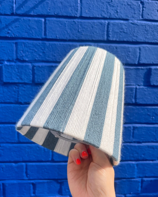 A hand holds a Slice Shade yarn lampshade featuring vertical stripes in light blue and white in front of a vibrant blue brick wall.
