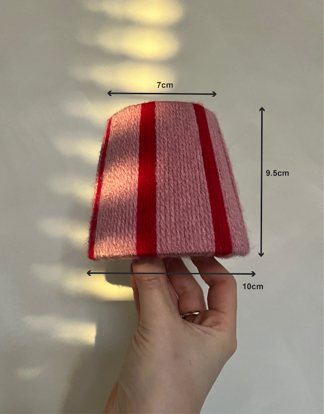 A hand holds a s Slice Shades yarn lampshade with pink and red vertical stripes. Black measurement arrows and labels indicate dimensions: 7 cm across the top, 10 cm across the base, and 9.