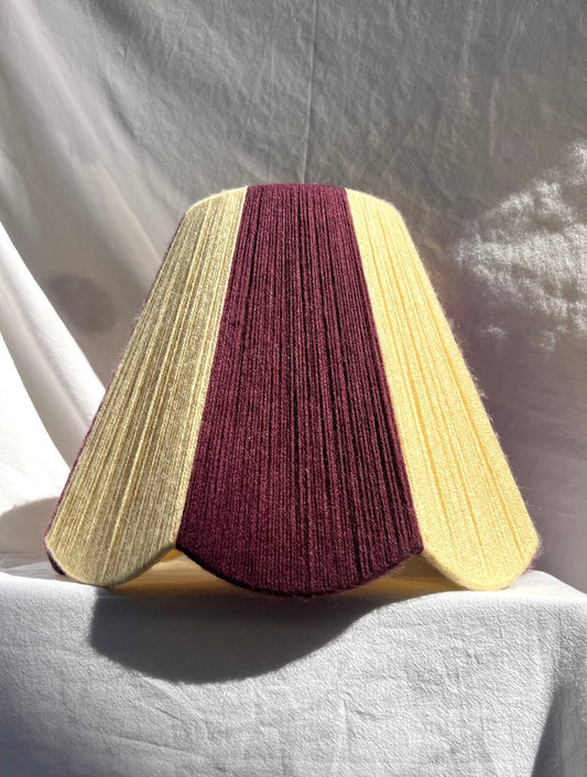 A Slice Shades lampshade with a handowoven yarn design of textured buttermilk yellow and deep burgundy, displayed on a draped white fabric background.