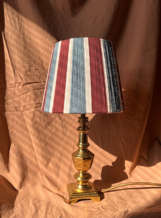 A table lamp with a Slice Shades yarn stripes in burgundy, blue, and white, mounted on a polished brass base, set against a draped brown fabric backdrop.