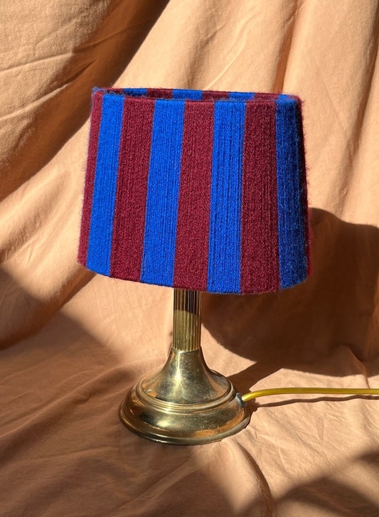 A table lamp with a striped yarn oval lampshade in alternating blue and burgundy stripes mounted on a metallic gold-toned base, set against a draped beige fabric backdrop.