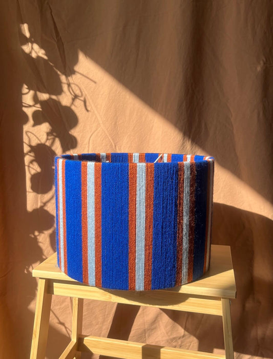 A Slice Shades yarn drum lampshade with vertical blue, orange, and white stripes, displayed on a small wooden stool against a draped brown fabric backdrop with leaf shadows.