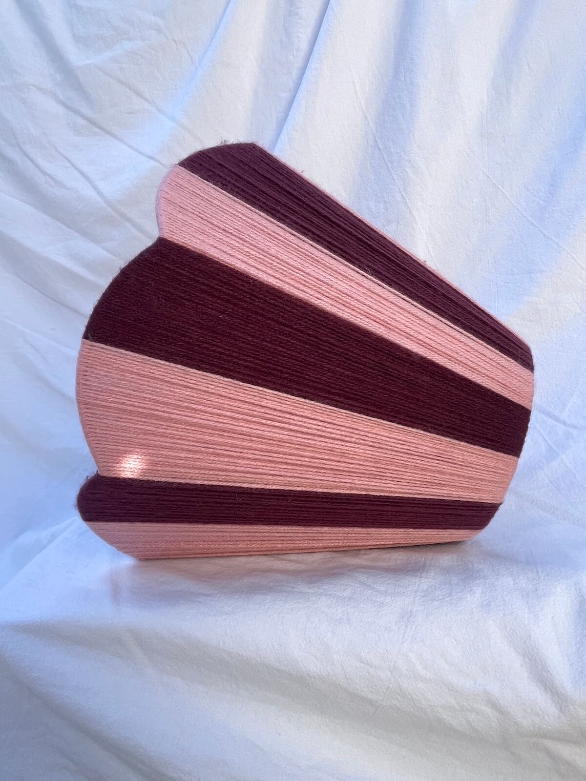A Slice Shades handwoven yarn lampshade with vertical stripes in dusty pink and deep burgundy, set against a draped white fabric background.