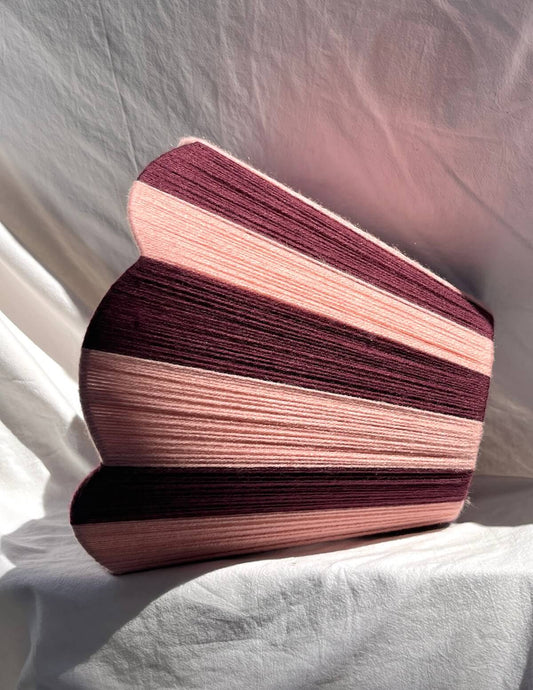 A Slice Shades handwoven yarn lampshade with vertical stripes in dusty pink and deep burgundy, set against a draped white fabric background.