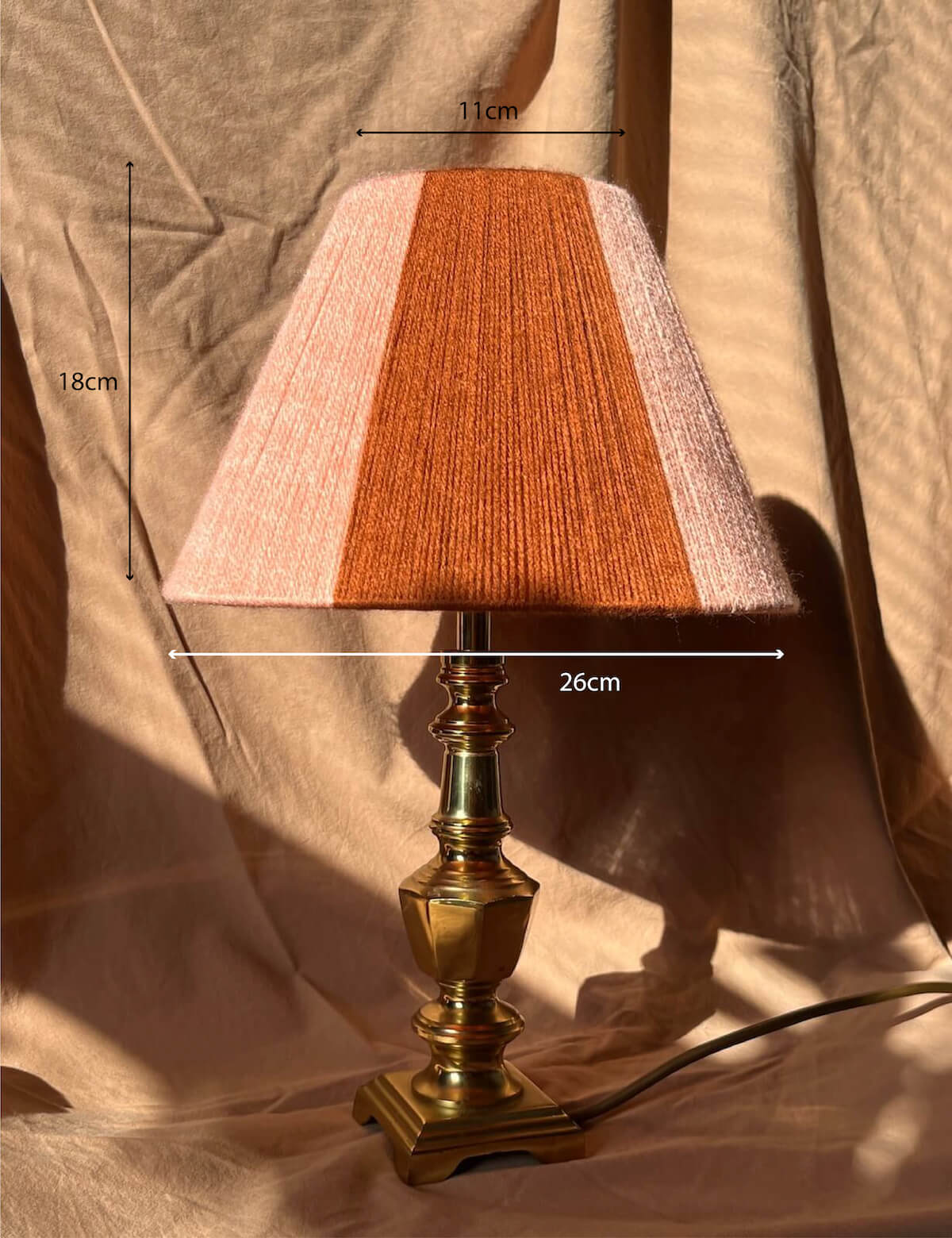 A Slice Shades table lamp with a pleated fabric shade in vertical stripes of light pink and rust orange, mounted on a polished brass base. The image features dimension overlays showing the shade's width (26cm), height (18cm), and top diameter (11cm) against a draped beige fabric backdrop.