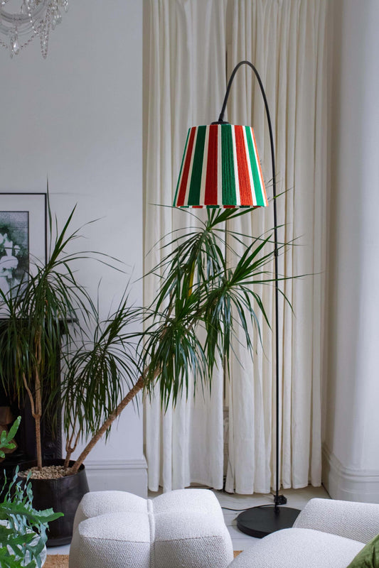 A tall black floor lamp with A Slice Shades handwoven yarn striped lampshade in Rust, Ecru & Jade Green standing in a modern living room next to a white sofa and potted plants.