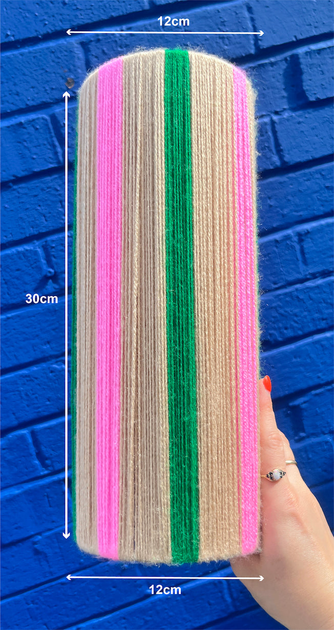 A hand holds a Slice Shades yarn lampshade covered in vertical stripes of pink, beige, and green yarn against a blue brick wall. White dimension lines with text show measurements of 30cm height, 12cm top width, and 12cm bottom width.