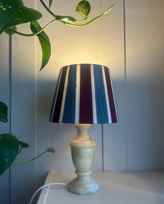 A Slice Shades yarn lampshade with a with vertical stripes of blue, burgundy, and cream, set on a white surface against a light gray panelled wall with a green plant leaf in the foreground.