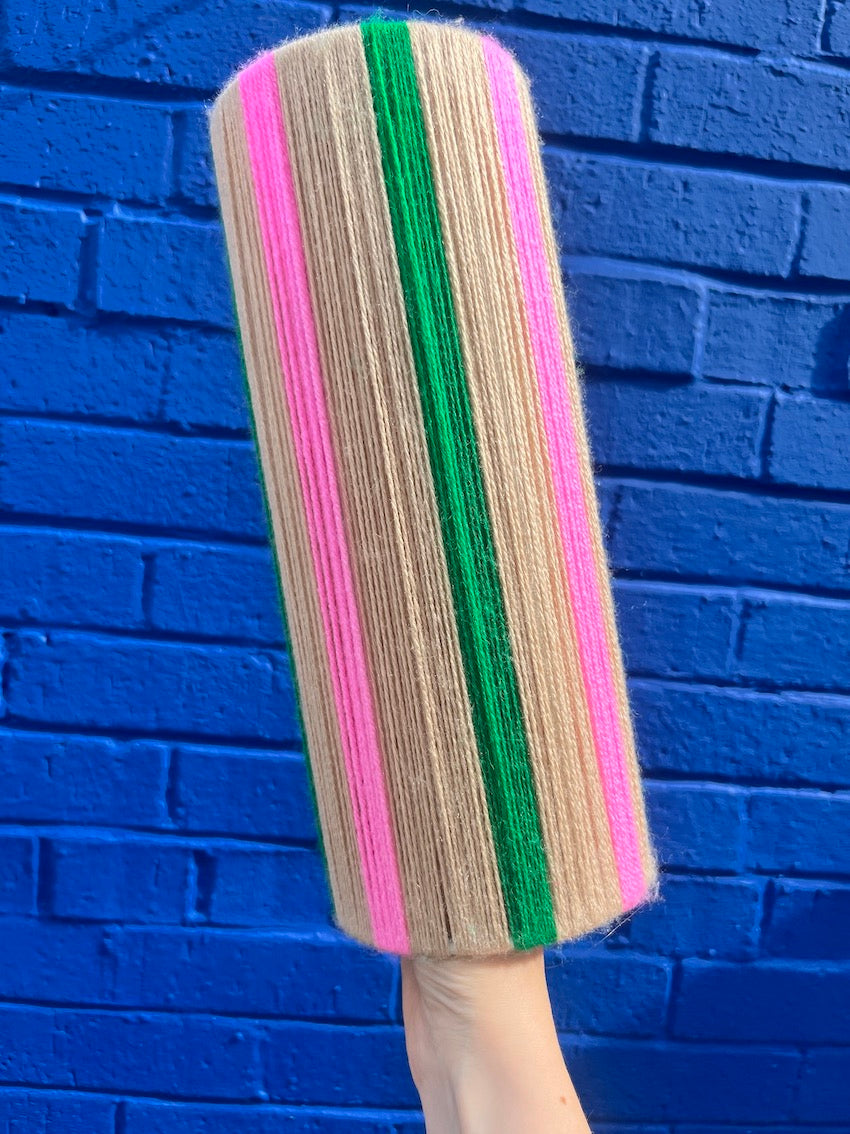 A Slice Shades lampshade featuring vertical stripes of beige, pink, and green yarn, held up against a bright blue brick wall background.