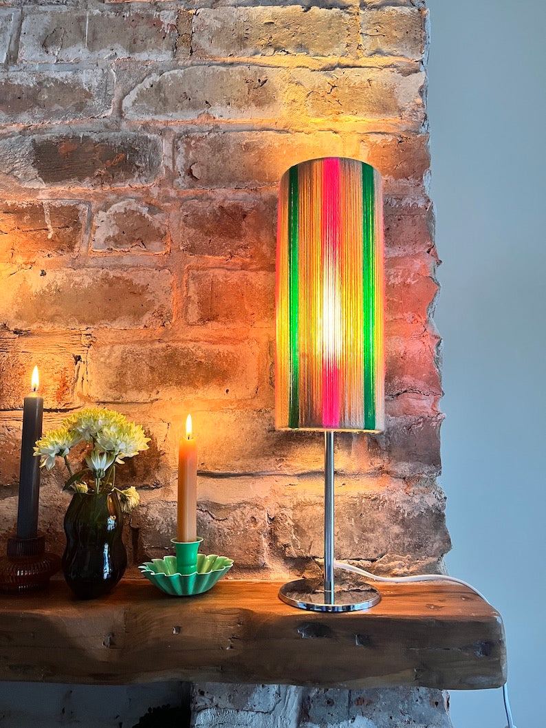 A Slice Shades yarn table lamp with a cylindrical shade featuring vertical multicoluor stripes in green, pink, orange,  standing on a silver metal base on a rustic wooden mantel against a brick wall.