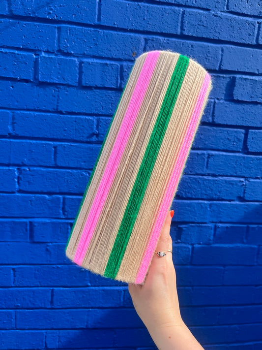 A hand holding a Slice Shades yarn lampshade with vertical pink, green, and beige stripes against a bright blue brick wall background.