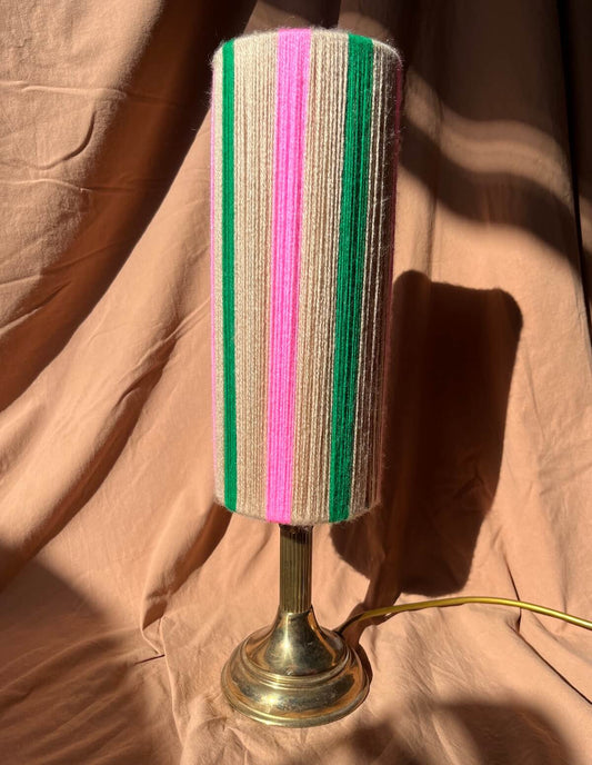 A vintage-style table lamp with a Slice Shades yarn lampshade featuring vertical stripes of beige, green, and pink, mounted on a brass-coloured metal base against a draped tan fabric backdrop.
