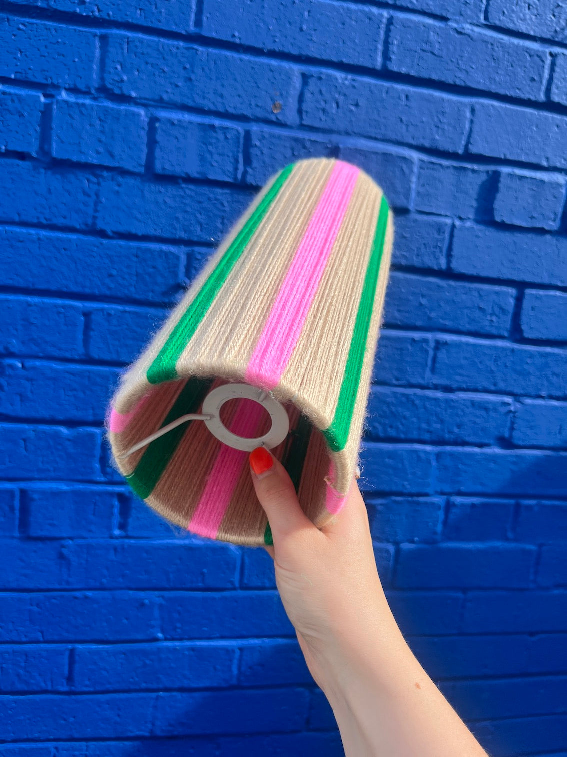 A hand holding a Slice Shades yarn lampshade with vertical pink, green, and beige stripes against a bright blue brick wall background.