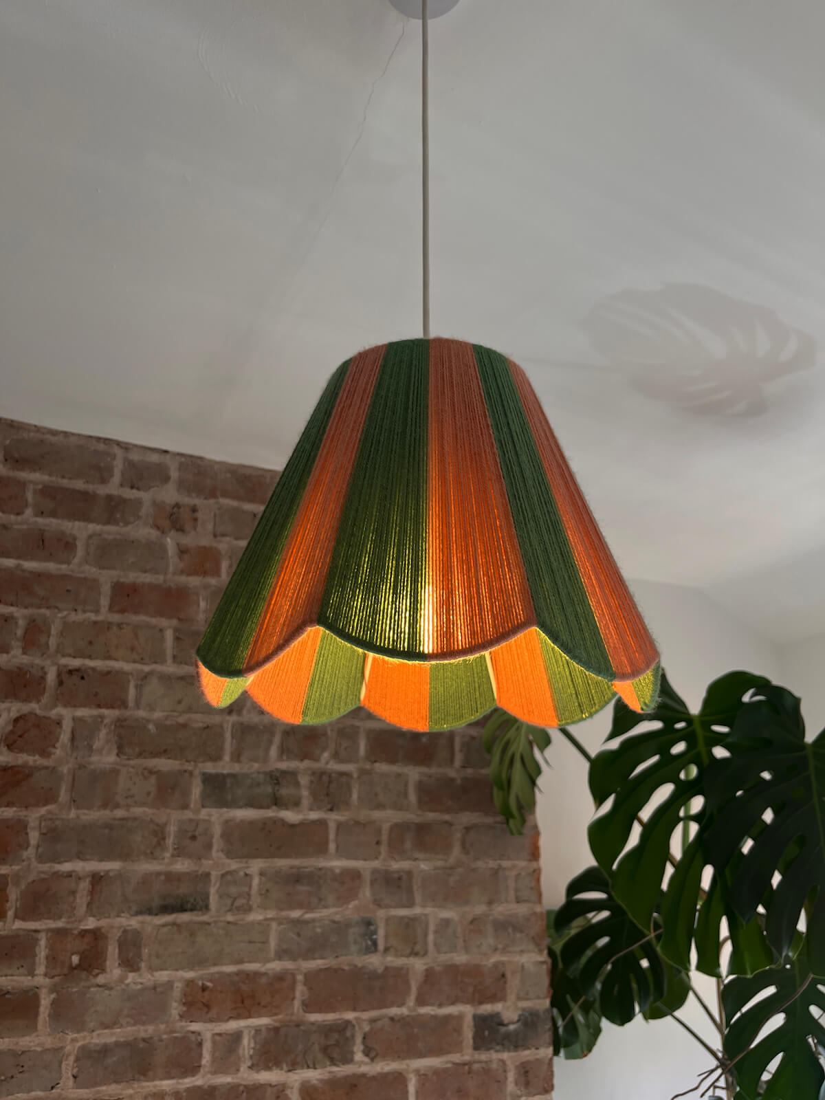A Slice Shades scallop yarn lampshade with vertical stripes of green and dusty pink lit up, hanging in a room with a brick wall and a potted monstera plant.