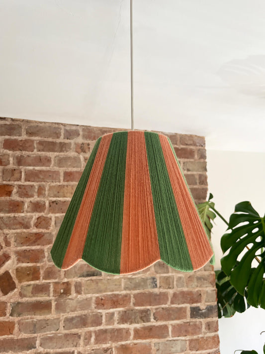 A Slice Shades scallop yarn lampshade with vertical stripes of green and dusty pink, hanging in a room with a brick wall and a potted monstera plant.
