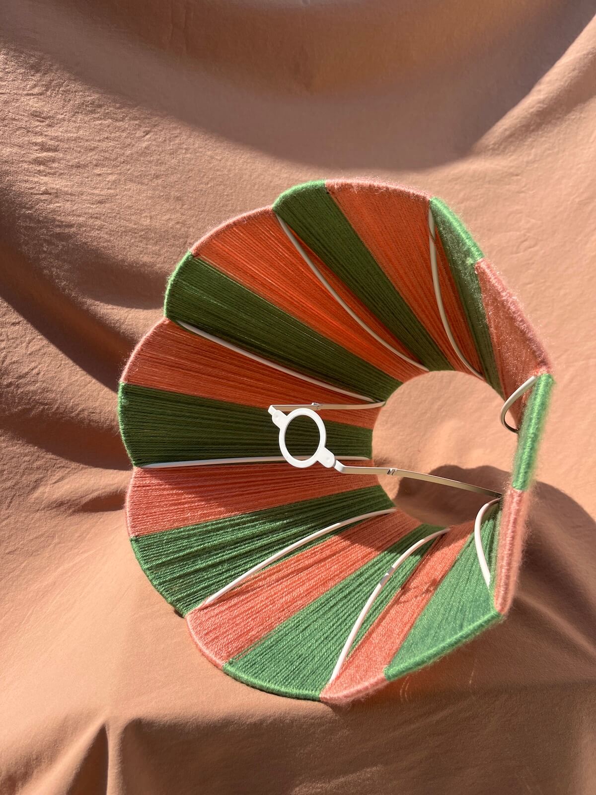 A Slice Shades scallop yarn lampshade  with striped segments of orange, green, and dusty pink thread, featuring a white circular bead and a silver metal ring, displayed on a draped beige fabric background.