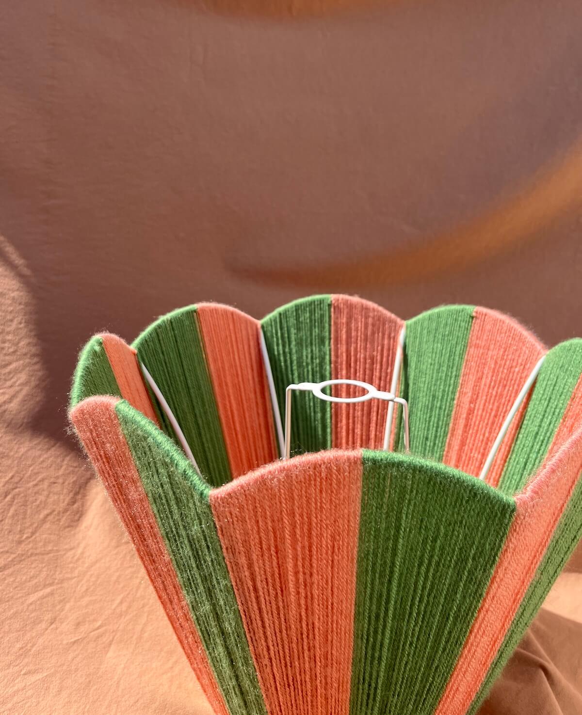 A close-up of a Slice Shades scallop yarn lampshade with alternating green and coral-pink vertical stripes, with a white wire support frame inside, set against a warm brown background.