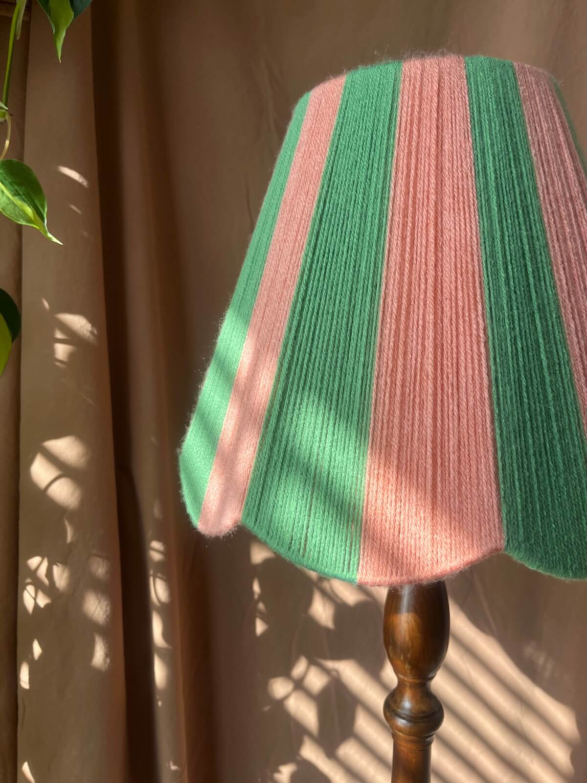 A Slice Shades yarn scalloped lampshade with vertical stripes of pea green and dusty pink, mounted on a dark wood lamp stand against a brown curtain backdrop with dappled sunlight and leaf shadows.
