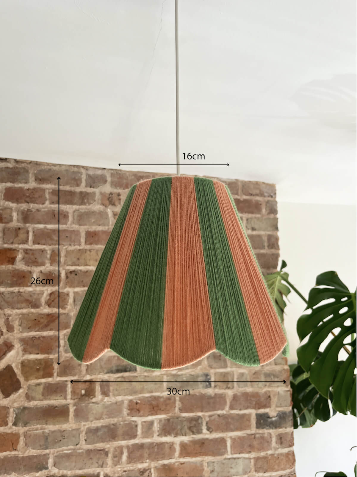 A Slice Shades scallop yarn lampshade with vertical stripes of green and dusty pink, hanging in a room with a brick wall and a potted monstera plant. Dimensions.