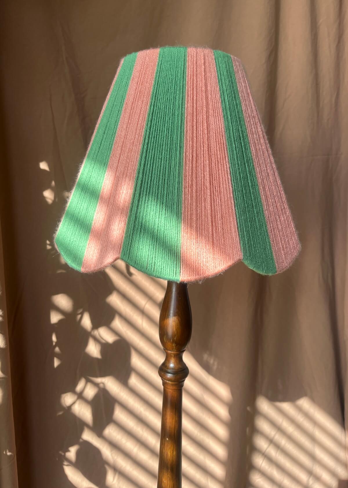 A Slice Shades scalloped yarn lampshade with vertical pink and green stripes, mounted on a dark brown wooden floor lamp stand against a beige curtain backdrop with dappled sunlight and leaf shadows.