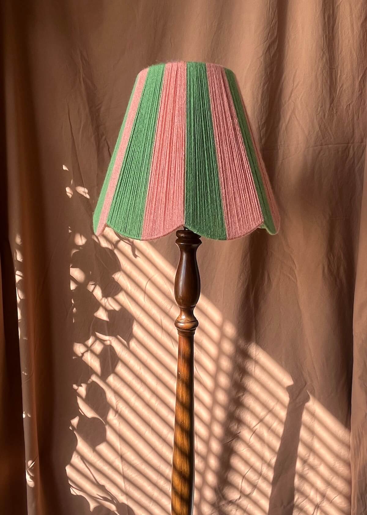 A vintage-style floor lamp with a Slice Shades scalloped yarn lampshade featuring alternating vertical stripes of pink and green, mounted on a twisted brown wooden pole, set against a draped brown fabric backdrop with window shadow patterns.