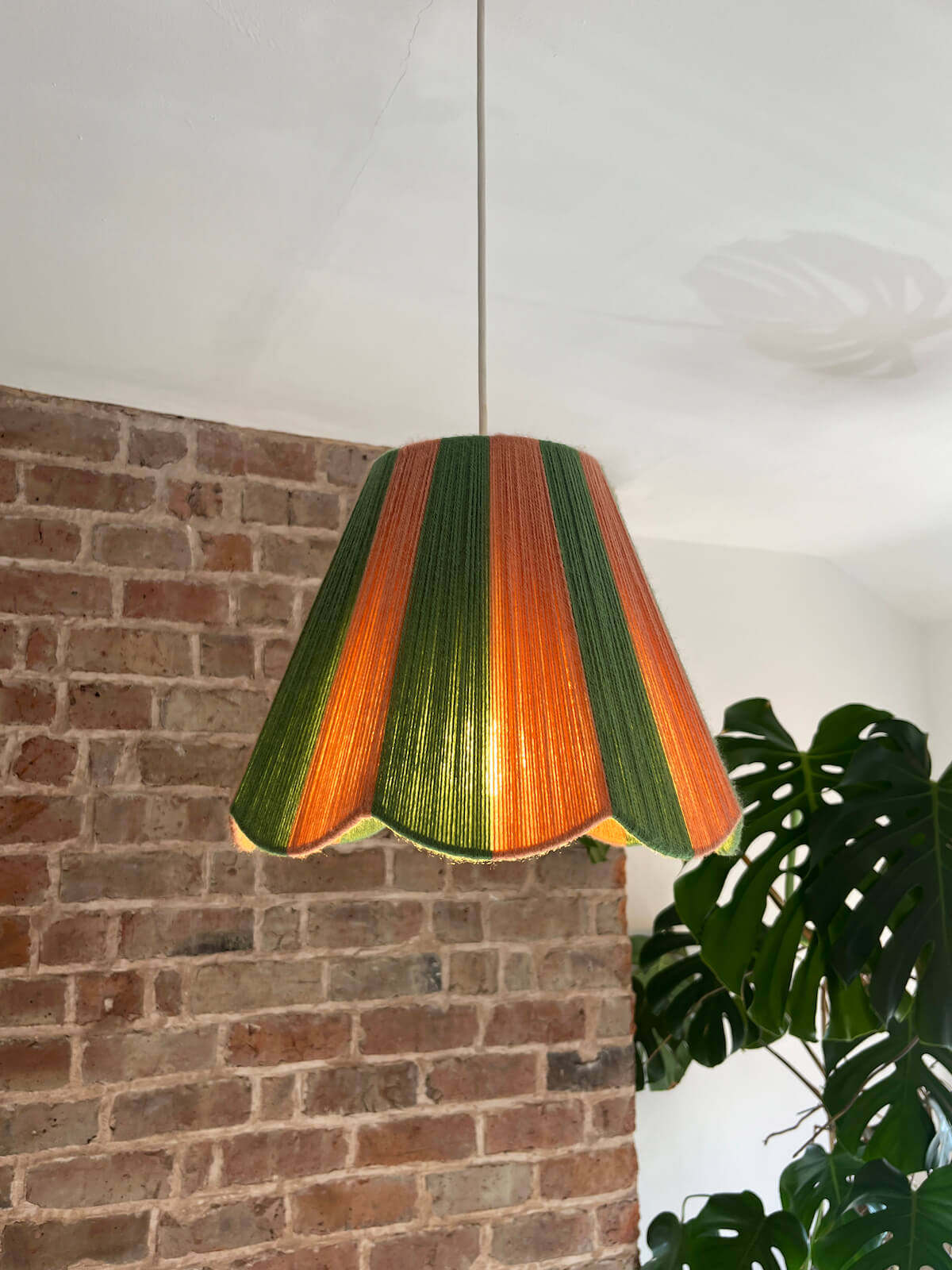 A Slice Shades scallop yarn lampshade with vertical stripes of green and dusty pink, hanging glowing in a room with a brick wall and a potted monstera plant.