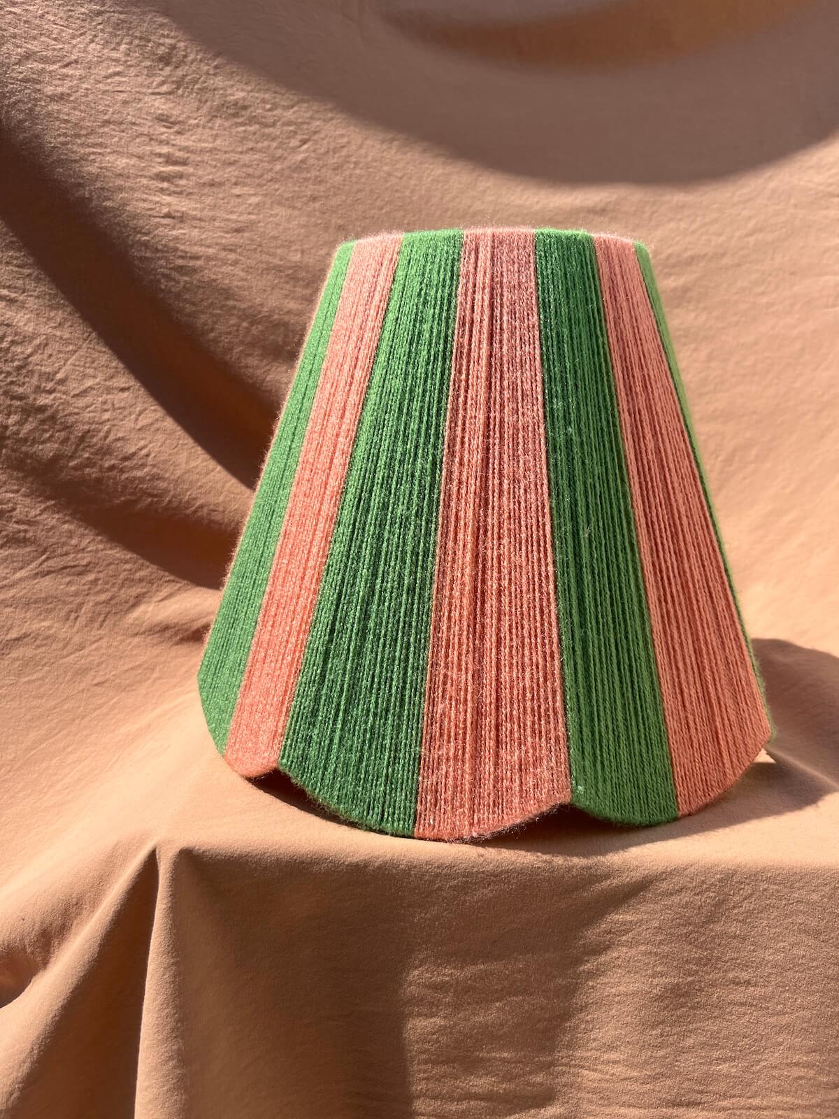 A Slice Shades scallop yarn lampshade with vertical stripes of green and dusty pink, displayed on draped beige fabric under warm sunlight.