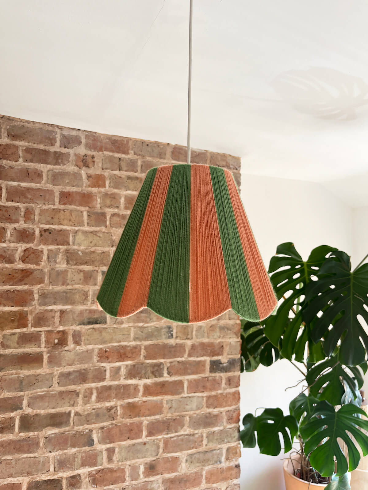 A Slice Shades scallop yarn lampshade with vertical stripes of green and dusty pink, hanging in a room with a brick wall and a potted monstera plant.