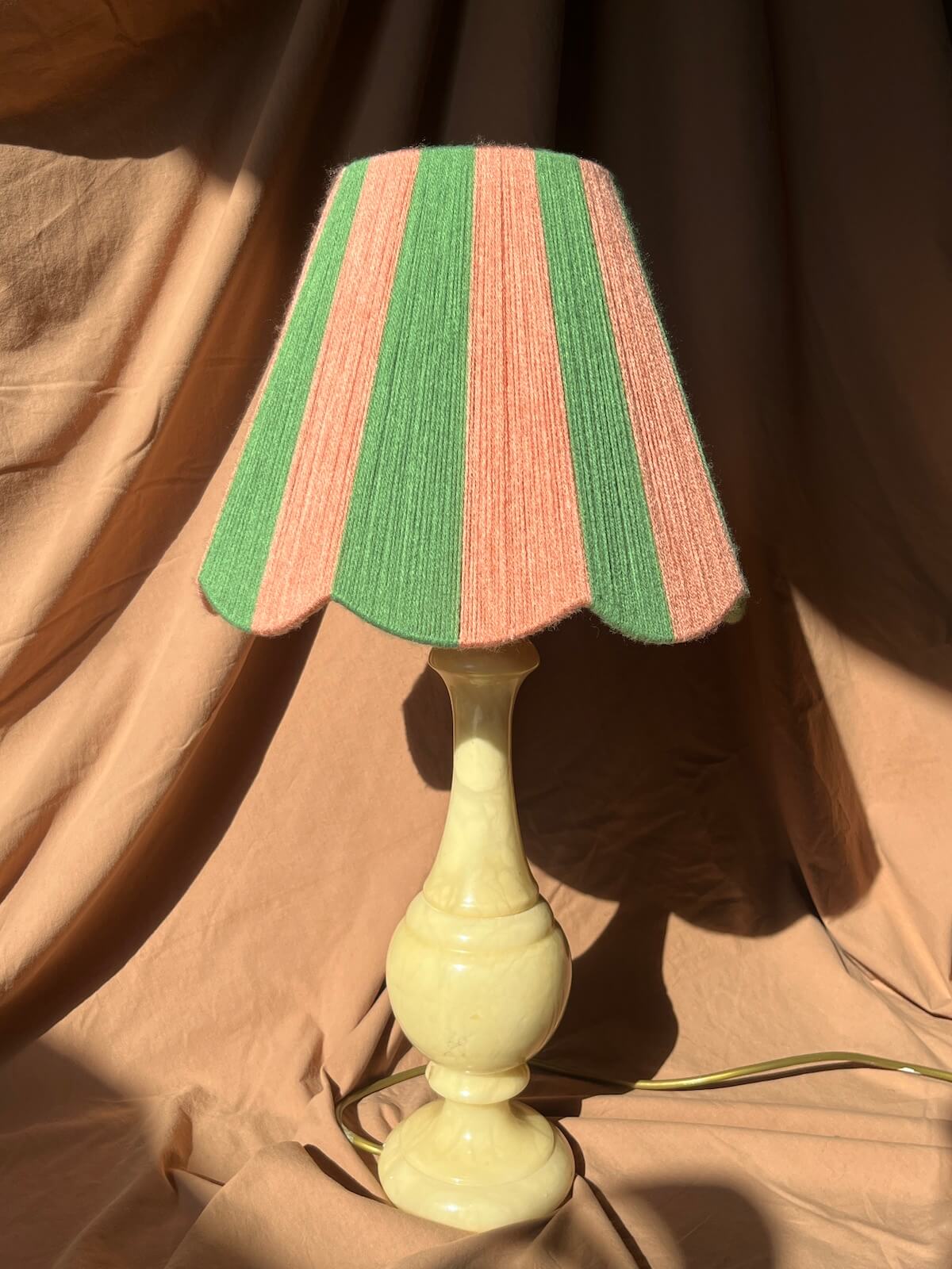 A vintage-style table lamp with a cream ceramic base and a Slice Shades scallop yarn lampshade featuring alternating vertical stripes of green and dusty pink, set against a draped tan fabric backdrop.