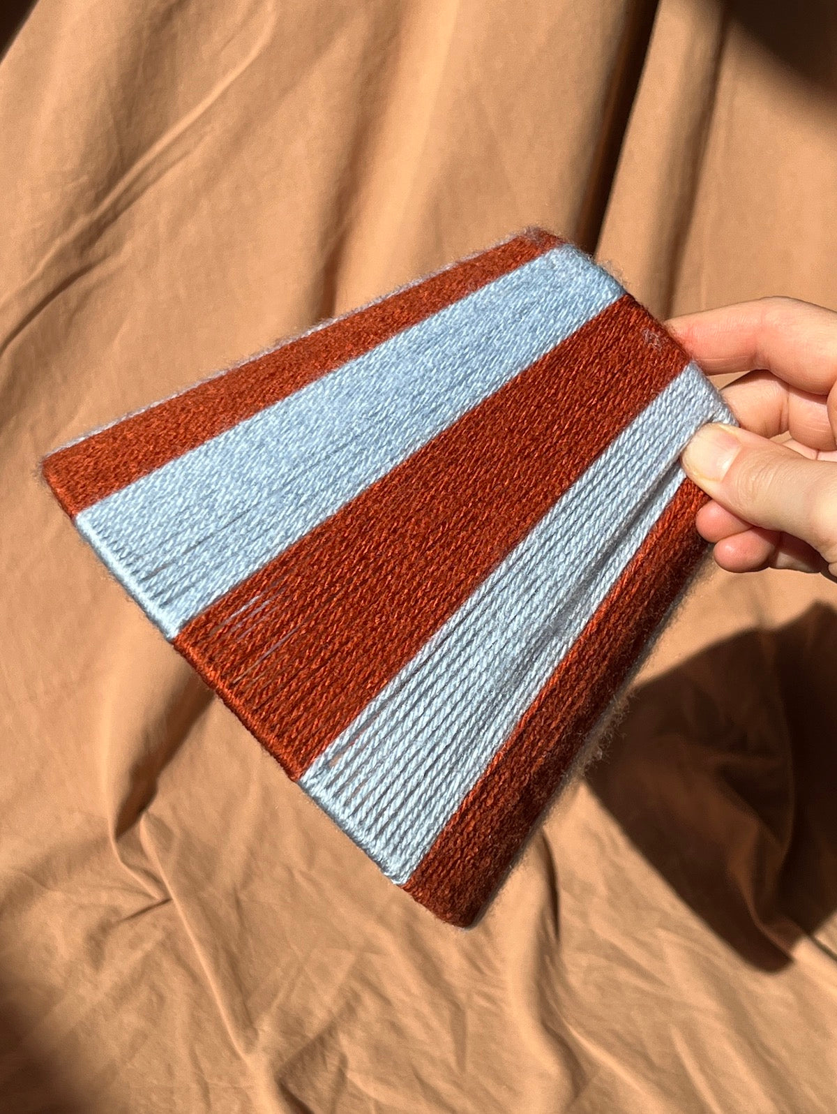 A hand holding a striped yarn string lampshade with alternating rust brown and light blue stripes, set against a draped tan fabric background.