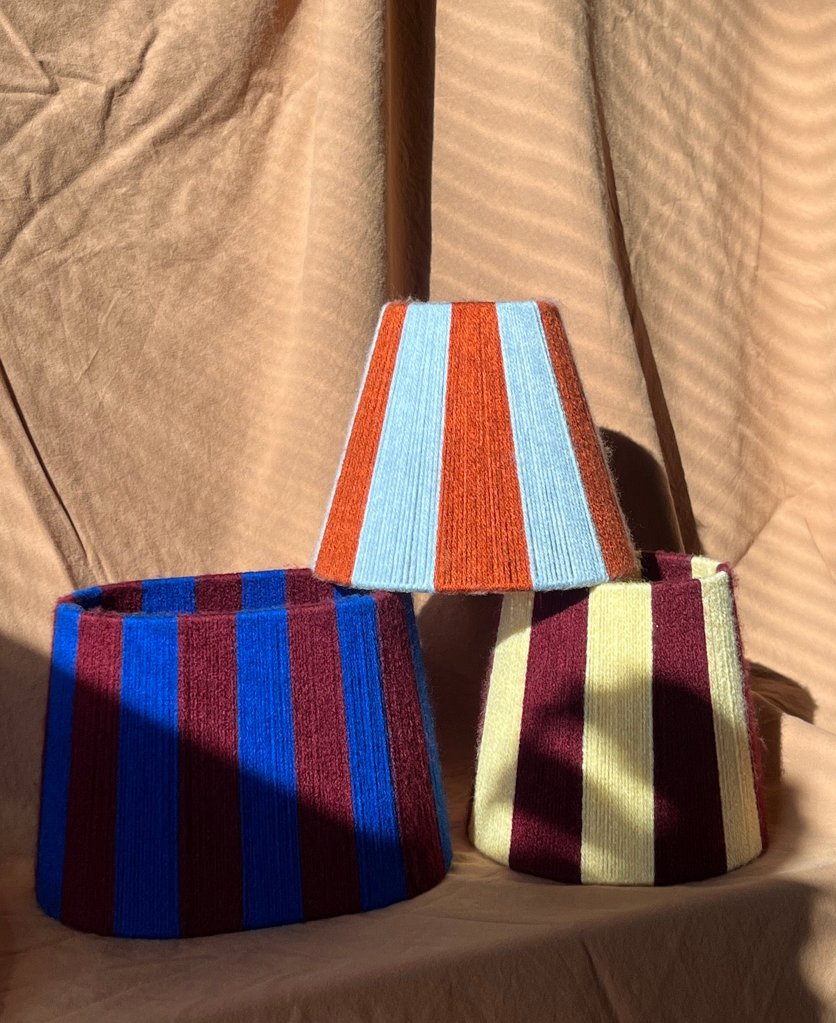 Three striped yarn lampshades with vertical stripes in different colourways (blue and burgundy, light blue, orange, and cream and burgundy) arranged on a beige fabric backdrop.