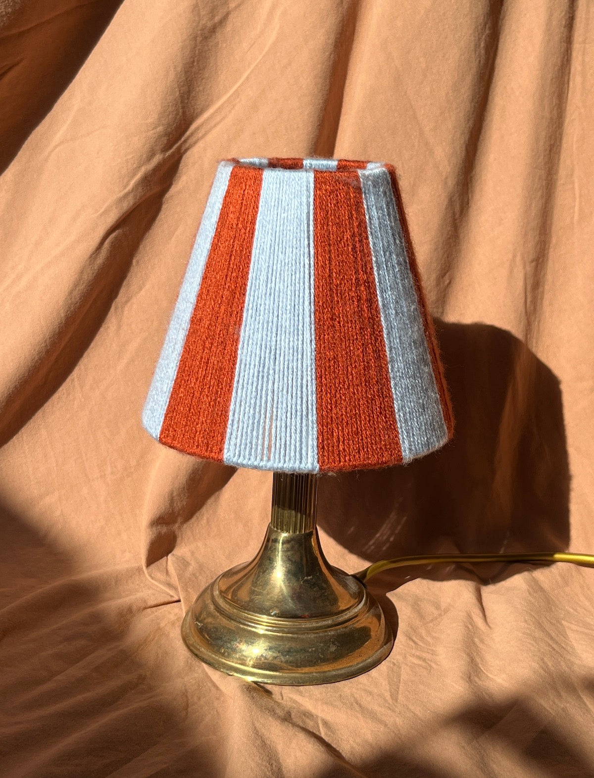 A small table lamp with a striped yarn shade in rust orange and light blue, mounted on a brass-coloured metal base, set against a draped tan fabric background.