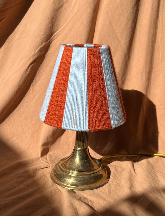 A small table lamp with a striped yarn shade in rust orange and light blue, mounted on a brass-coloured metal base, set against a draped tan fabric background.