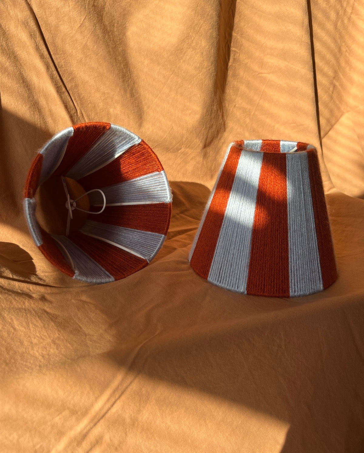Two string yarn lampshades with vertical rust orange and light blue stripes, displayed on draped tan fabric under direct sunlight.