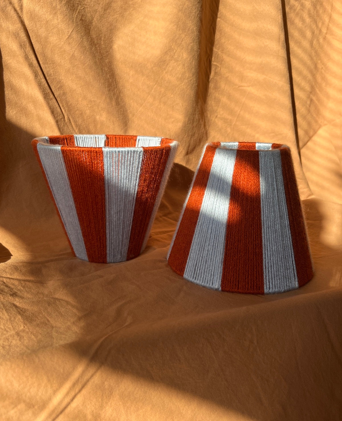 Two handmade string yarn lampshades and light blue and rust orange stripes, placed on a draped tan fabric background with strong sunlight and shadows.