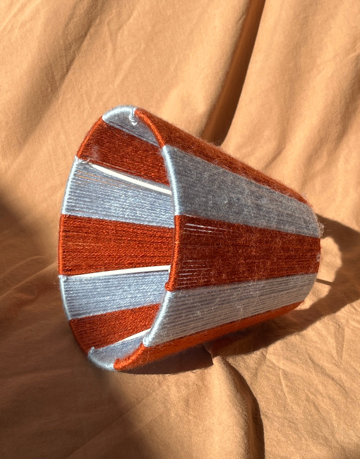 A handmade string yarn lampshade with alternating rust orange and light blue stripes, photographed on a draped tan fabric background.