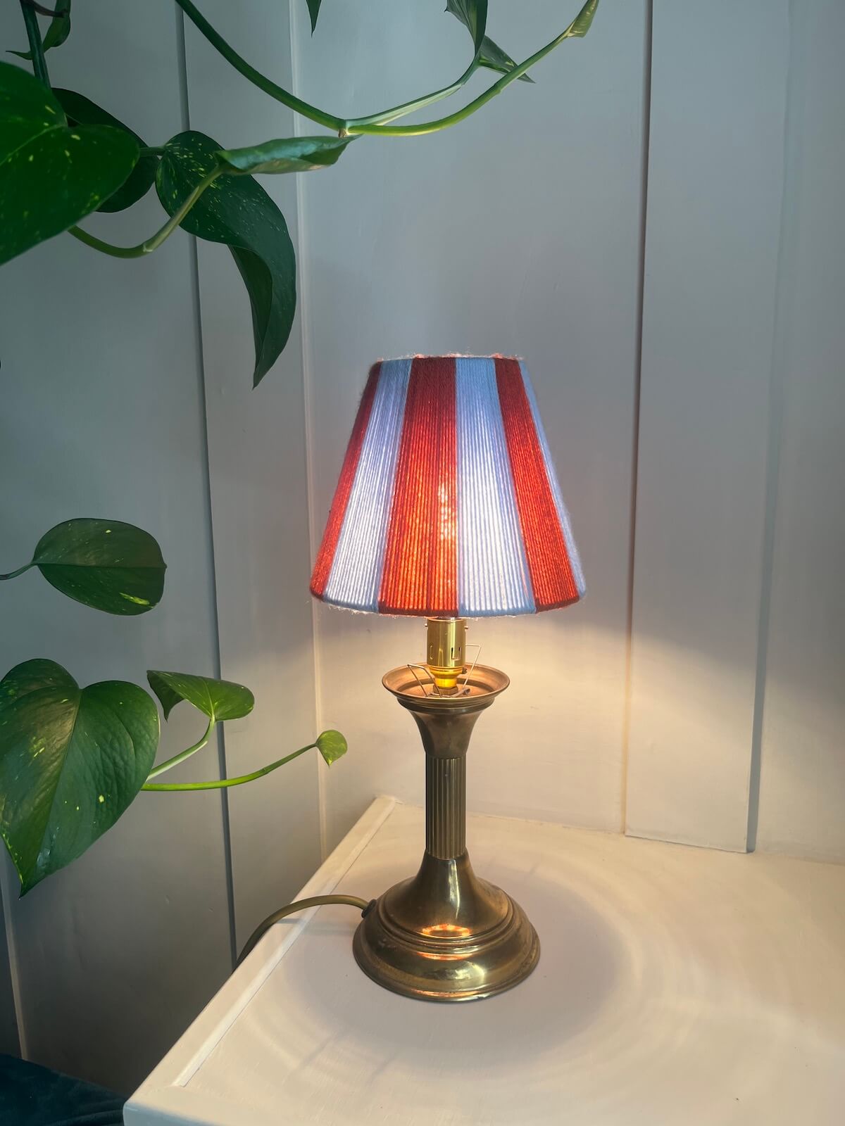 A Slice Shades yarn lampshade in rust and blue stripes on a brass-coloured metal base, illuminated and placed on a white surface against a white panelled wall with green plant leaves in the foreground.