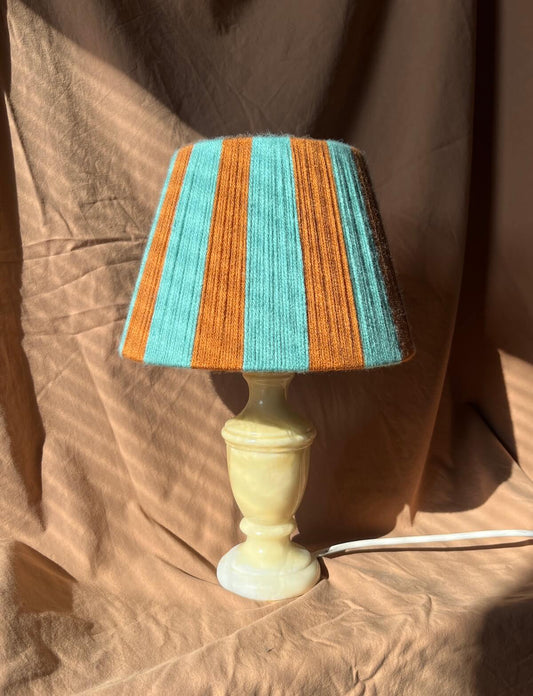 A small table lamp with a Slice Shades yarn lampshade featuring alternating turquoise blue and rust-orange vertical stripes, set on a pale cream-coloured pedestal base against a draped brown fabric backdrop.