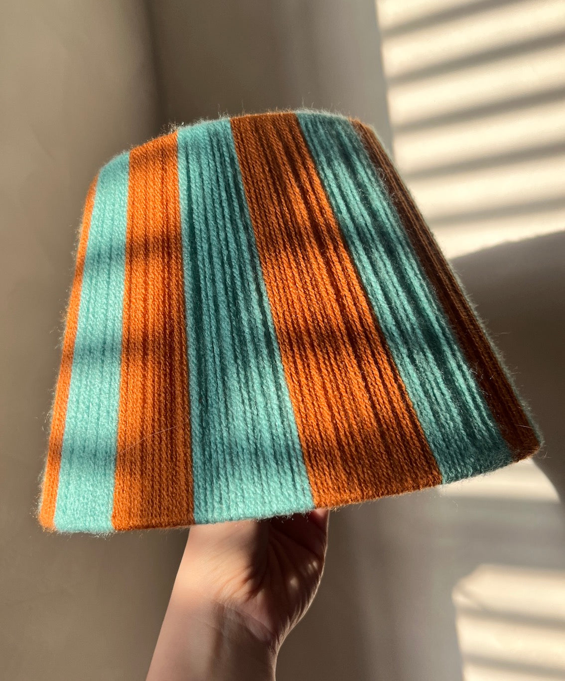 A hand holds a Slice Shades lampshade with vertical stripes in burnt orange and turquoise lit by warm sunlight with soft window shadow patterns.