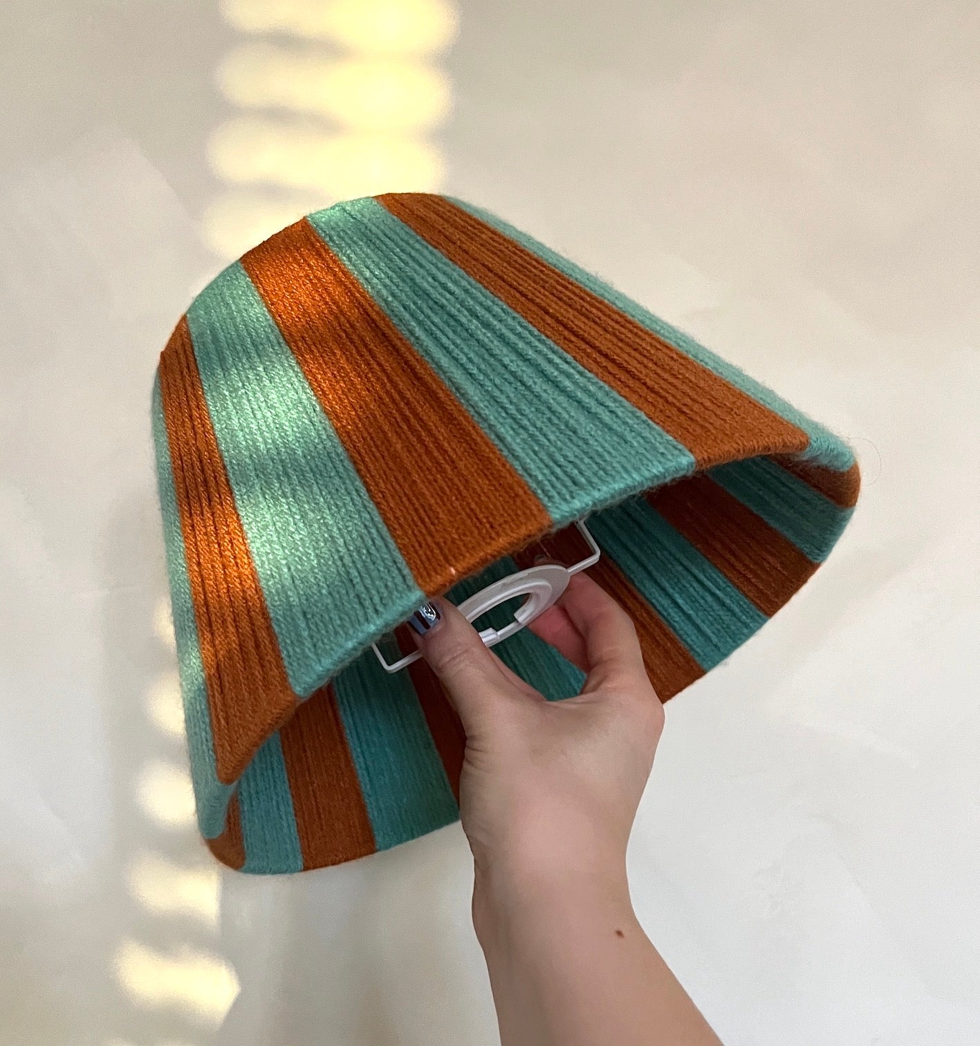 A hand holding a Slice Shades yarn lampshade with alternating turquoise and rust orange stripes, photographed on a light neutral surface with soft window-light shadows.