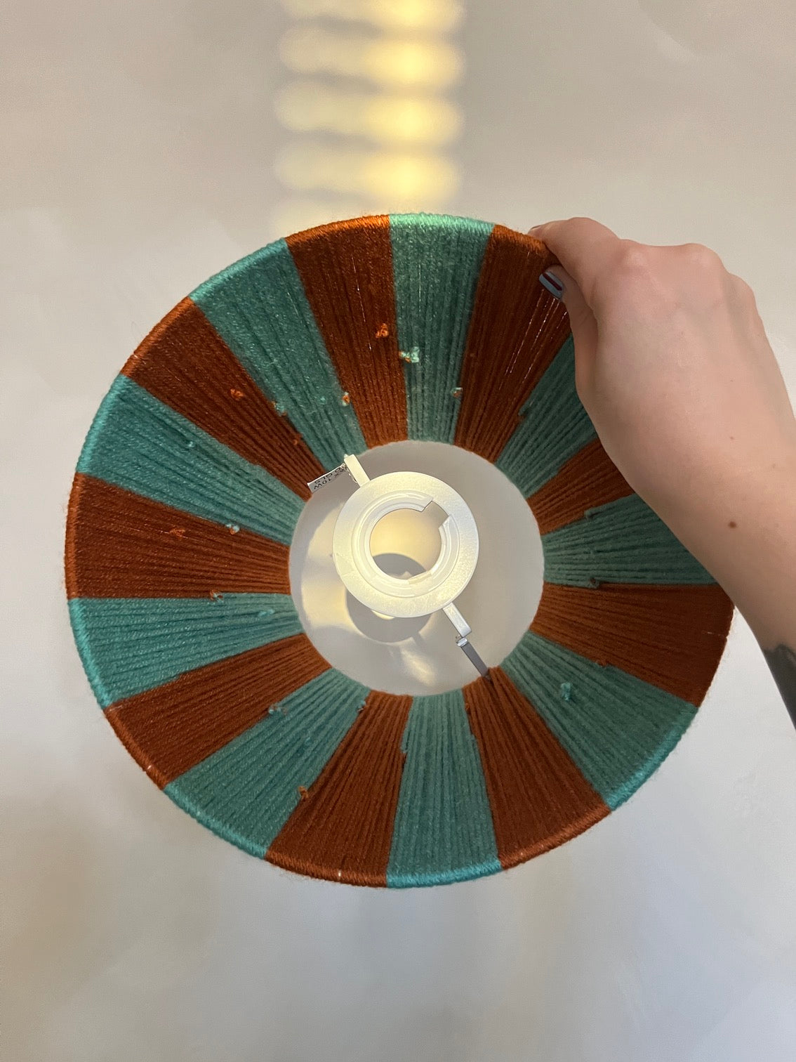 A hand holding a Slice Shadesyarn woven lampshade with alternating teal and rust-brown radial stripes, photographed against a softly blurred indoor background.
