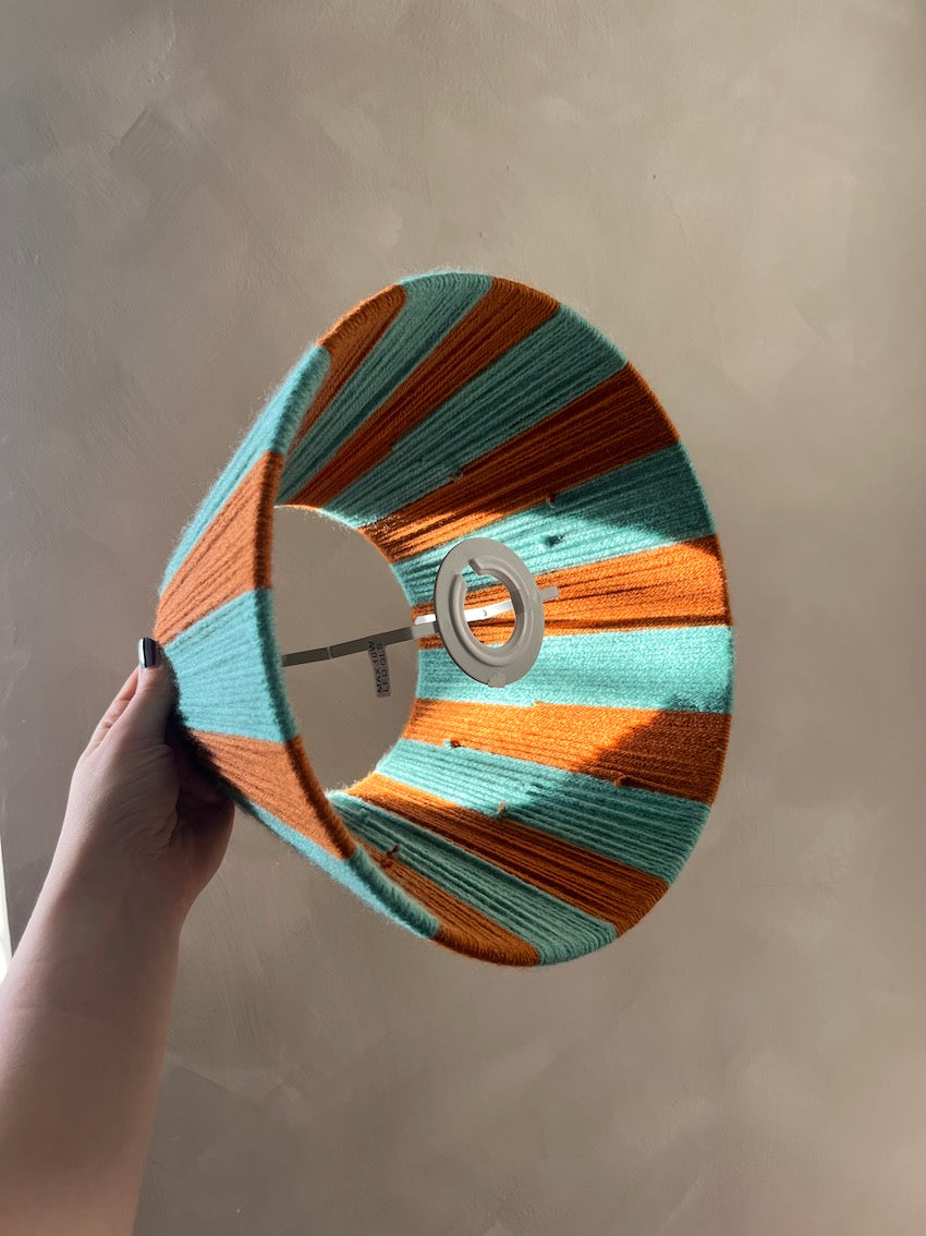 A hand holding a Slice Shades yarn woven lampshade with alternating stripes of turquoise and burnt orange, set against a textured beige wall