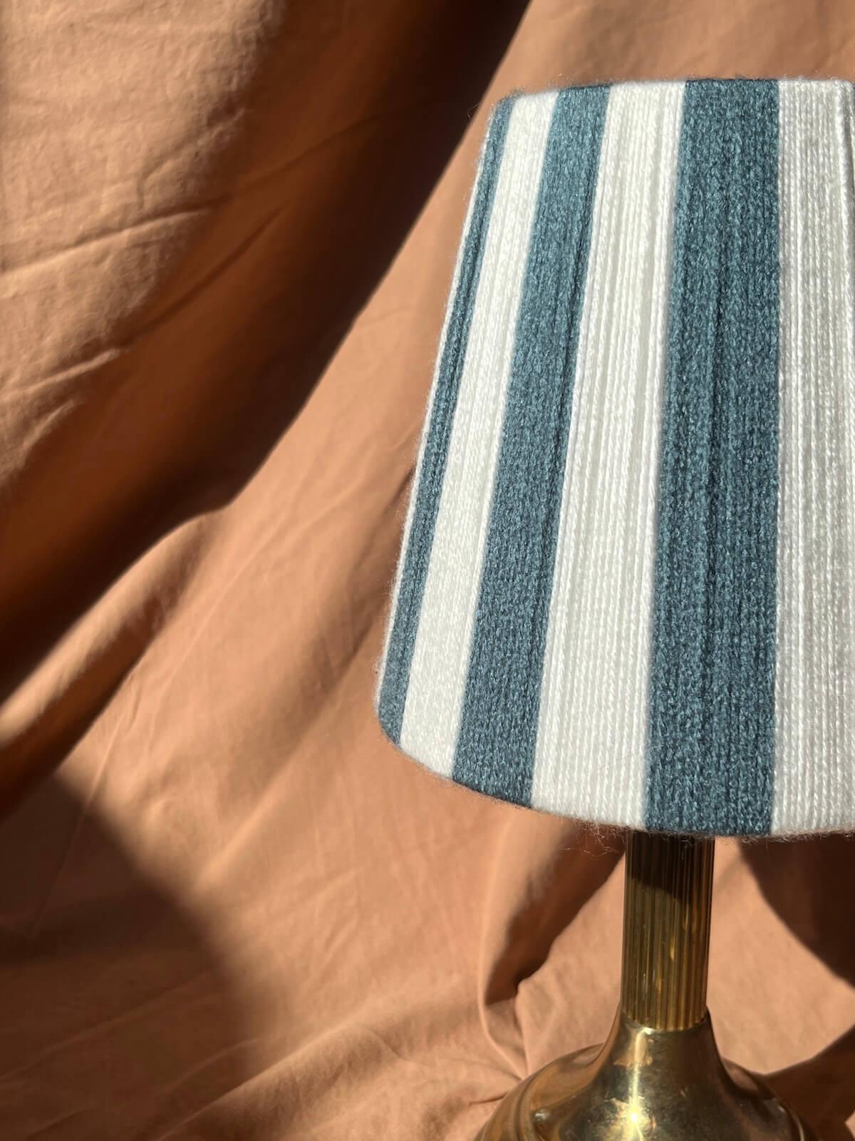 A close-up of a Slice Shades table lamp shade with vertical blue-gray and off-white stripes, mounted on a brass-coloured base against a draped peach fabric background.