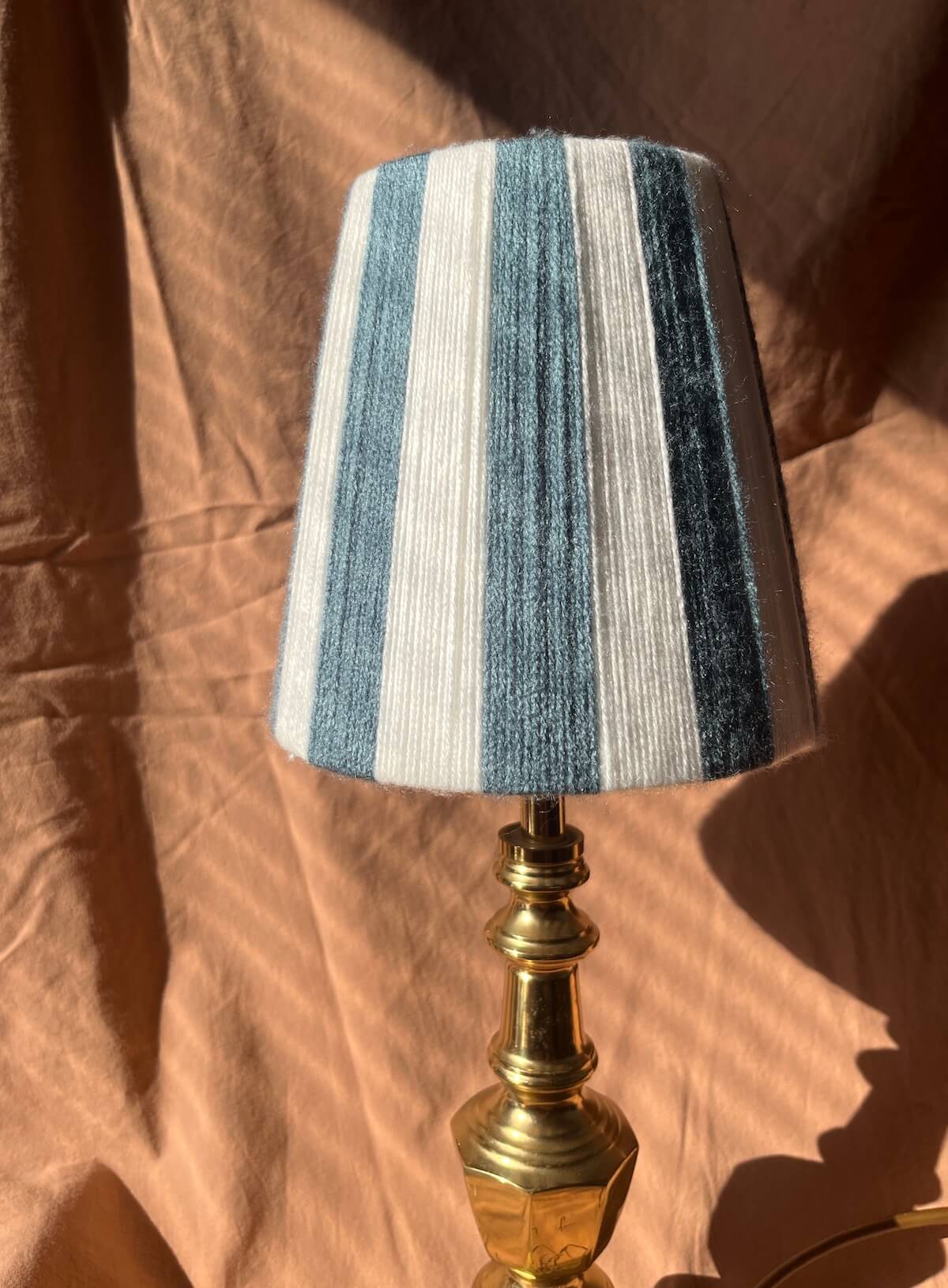 A Slice Shades table lamp with a blue and white vertically striped fabric shade and a polished brass base, set against a draped brown fabric background.