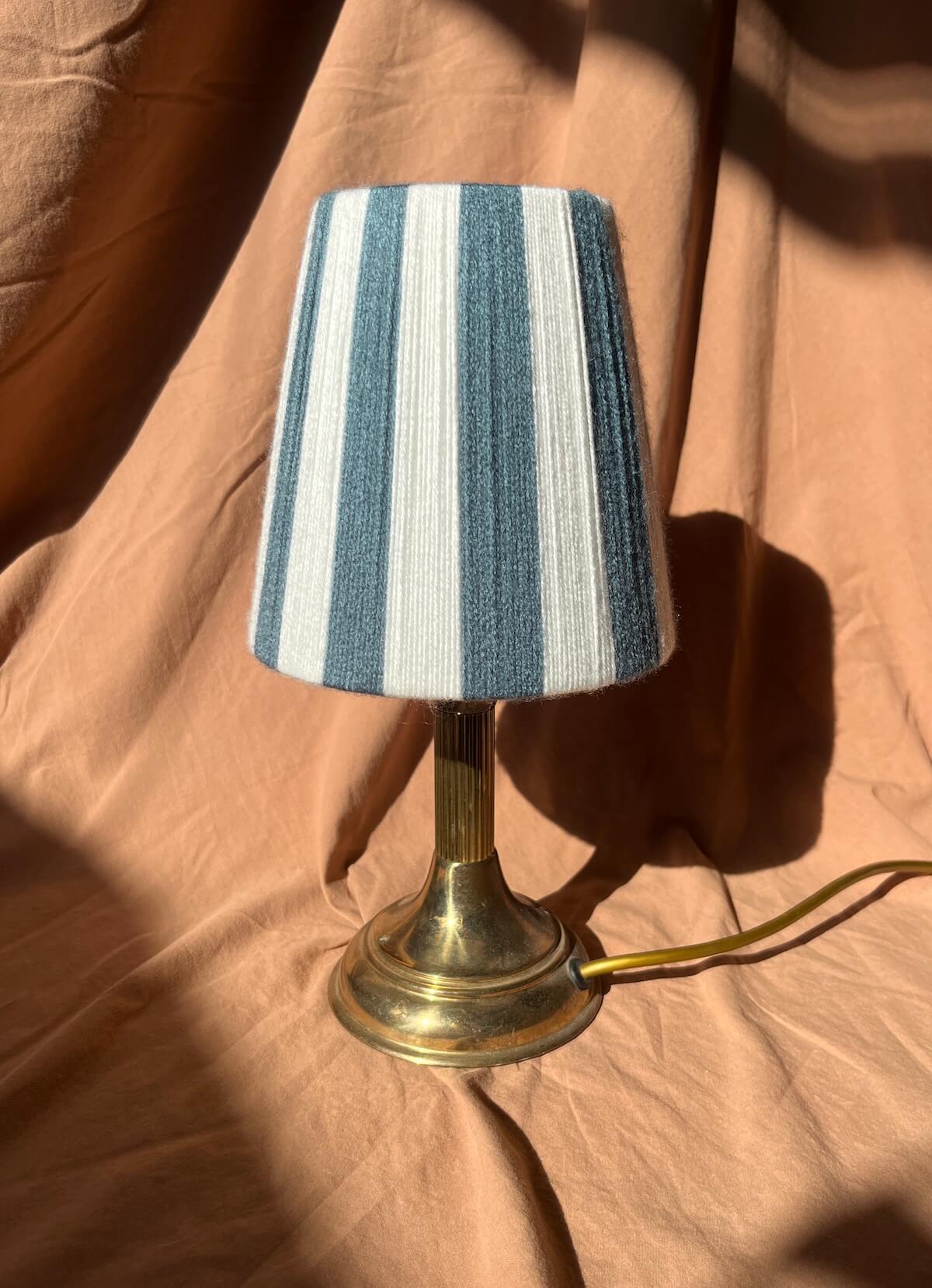 A small table lamp with a blue and white vertically striped yarn Slice Shade shade and a brass-colored metal base, set against a draped peach fabric background.