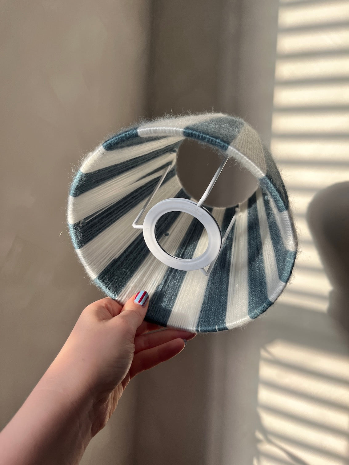 A hand holding a Slice Shades lampshade with a blue and white striped pattern, set against a background of beige with soft window shadow pattern