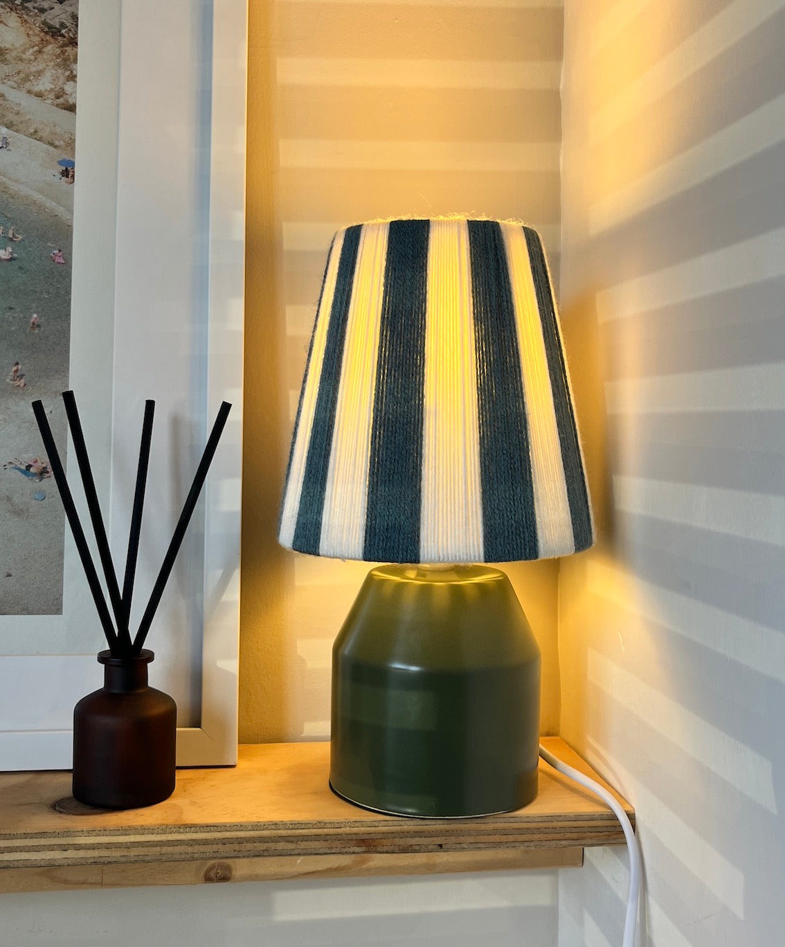 A table lamp with a dark green ceramic base and a Slice Shades yarn lampshade featuring vertical blue and ecrus stripes, illuminated with a warm yellow glow. It is styled on a wooden shelf next to a reed diffuser against a striped wallpaper background.