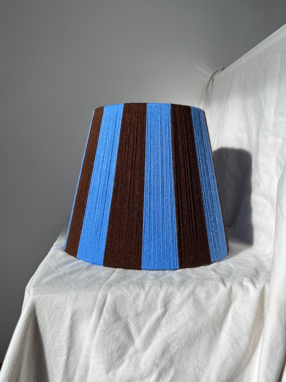 A Slice Shades handmade yarn striped lampshade with alternating vertical stripes of blue and dark brown, displayed on a white fabric backdrop.

