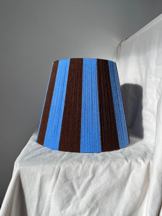 A Slice Shades handmade yarn striped lampshade with alternating vertical stripes of blue and dark brown, displayed on a white fabric backdrop.
